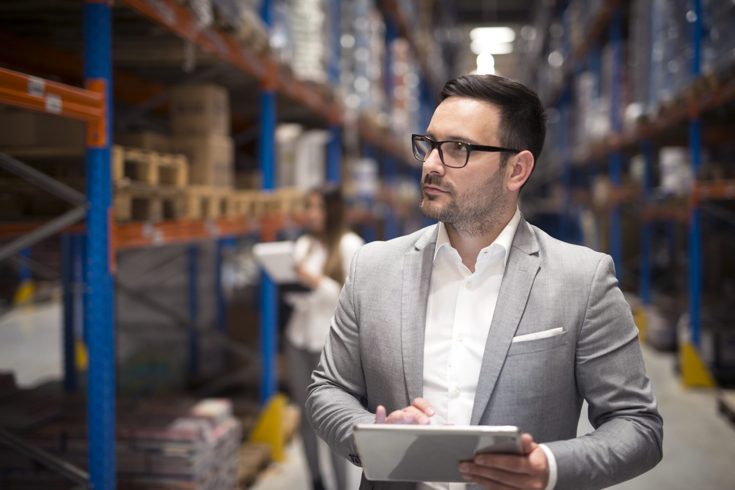 Portrait of successful businessman manager CEO holding tablet and walking through warehouse storage area looking towards shelves.